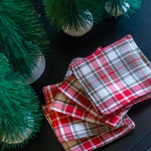 Peut inclure: Quatre dessous de verre carrés en tissu à motif écossais rouge, blanc et gris. Les dessous de verre sont empilés sur une surface noire, avec deux petits sapins verts en arrière-plan.
