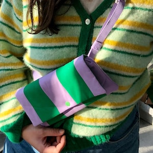 May include: A purple and green striped fabric bag with a purple strap. The bag is worn over the shoulder of a person wearing a green and yellow striped cardigan.