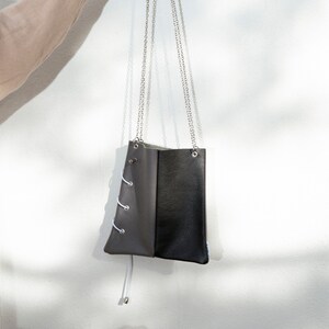 Könnte beinhalten: Zweifarbiges Lederhandtasche mit silberner Kette. Die Tasche ist halb grau und halb schwarz, mit weißen Schnürdetails auf der grauen Seite. Die Tasche wird von einer silbernen Kette gehalten.