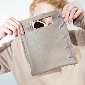 Könnte beinhalten: Eine graue Ledertasche mit einem Ausschnitt für Sonnenbrillen und einem rosa-weißen Kordelverschluss.