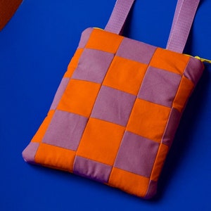 May include: A purple and orange checkered crossbody bag with a purple strap. The bag is made of fabric and has a zipper closure.
