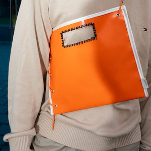 Könnte beinhalten: Eine leuchtend orangefarbene, rechteckige Tasche mit weißem Besatz und einem kleinen, rechteckigen Fenster. Die Tasche hat ein weißes Schnürdetail an der Seite und wird über einem beigefarbenen Pullover getragen.