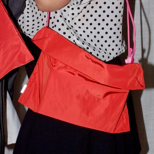 May include: A vibrant red shoulder bag with a pink strap. The bag has a rectangular shape and a folded-over top. The person in the image is wearing a white blouse with black polka dots and a black skirt.