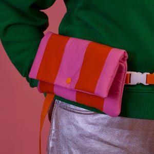 Könnte beinhalten: Eine rosa-orange gestreifte Bauchtasche mit einem gelben und orangefarbenen geflochtenen Gürtel. Die Bauchtasche wird über einem grünen Sweatshirt und einer silbernen Hose getragen.