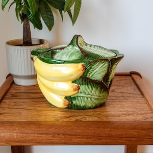 Peut inclure: Un bol en céramique avec couvercle en forme de grappe de bananes jaunes nichées parmi des feuilles vertes texturées. Le couvercle présente un motif de feuille sculptée formant une poignée, avec des accents bruns sur les bords des feuilles.