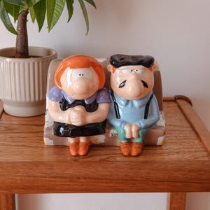 Peut inclure: Une figurine en céramique représentant deux personnages de dessin animé assis sur un banc. Le personnage de gauche a des cheveux orange, un haut violet et une jupe noire. Celui de droite porte un béret noir, une chemise bleu clair et un pantalon vert.