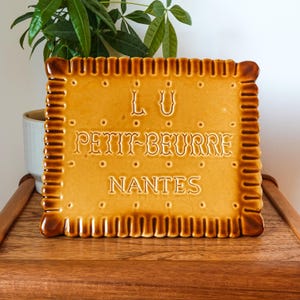 Peut inclure: Une plaque décorative en céramique carrée en forme de biscuit LU Petit-Beurre avec une finition émaillée brun doré. La surface présente le texte en relief LU, PETIT-BEURRE et NANTES, entouré d'un bord festonné et de petites indentations.