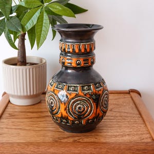 Peut inclure: Un vase en céramique présentant une glaçure brun foncé avec des motifs en relief orange vif et crème. Le design comprend des bandes horizontales de formes géométriques et de motifs circulaires autour du col et du corps arrondi du récipient.