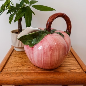Op de afbeelding: Een keramische kan in de vorm van een perzik met een glanzende roze afwerking, voorzien van een donkerbruin gebogen handvat en een groen bladaccent bij de tuit. De binnenkant van de kan is wit en de buitenkant heeft subtiele verticale penseelstreken.