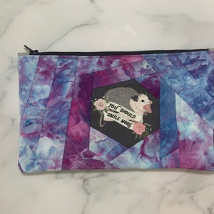 May include: A zippered pouch with a geometric pattern in shades of purple and blue. The pouch features a graphic of an opossum with the text "YOU SHOULD SMILE MORE" on a banner, surrounded by pink roses.