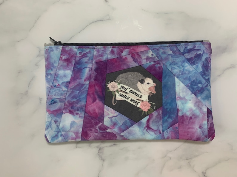 May include: A zippered pouch with a geometric pattern in shades of purple and blue. The pouch features a graphic of an opossum with the text "YOU SHOULD SMILE MORE" on a banner, surrounded by pink roses.