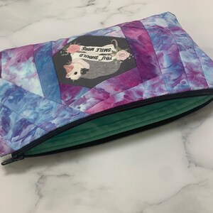 May include: A quilted zippered pouch with a vibrant tie-dye pattern in shades of blue, purple, and pink. The pouch features a central patch with a possum illustration and the text "YOU SHOULD SMILE MORE". The interior lining is a soft green.