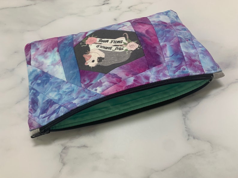 May include: A quilted zippered pouch with a vibrant tie-dye pattern in shades of blue, purple, and pink. The pouch features a central patch with a possum illustration and the text "YOU SHOULD SMILE MORE". The interior lining is a soft green.