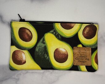 May include: A rectangular zippered pouch with a black background and a pattern of avocado halves. The avocados are green and yellow with brown seeds. A small leather tag with the text "Curious Dog Designs" is sewn on the front.