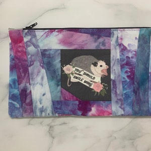 May include: A rectangular zippered pouch with a tie-dye pattern in shades of blue, purple, and pink. The center features a graphic of an opossum with the text "YOU SHOULD SMILE MORE" on a banner. The pouch is likely for cosmetics or small items.