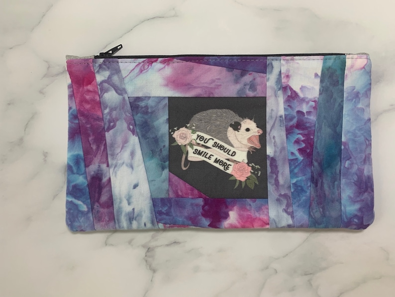 May include: A rectangular zippered pouch with a tie-dye pattern in shades of blue, purple, and pink. The center features a graphic of an opossum with the text "YOU SHOULD SMILE MORE" on a banner. The pouch is likely for cosmetics or small items.