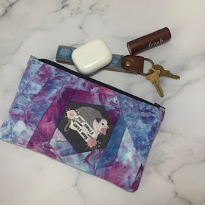 May include: A quilted fabric pouch with a black zipper, featuring a tie-dye pattern in shades of blue, purple, and pink. The pouch has an image of an opossum with the text "YOU SHOULD SMILE MORE". Beside the pouch are keys, a lipstick, and a pair of white wireless earbuds.