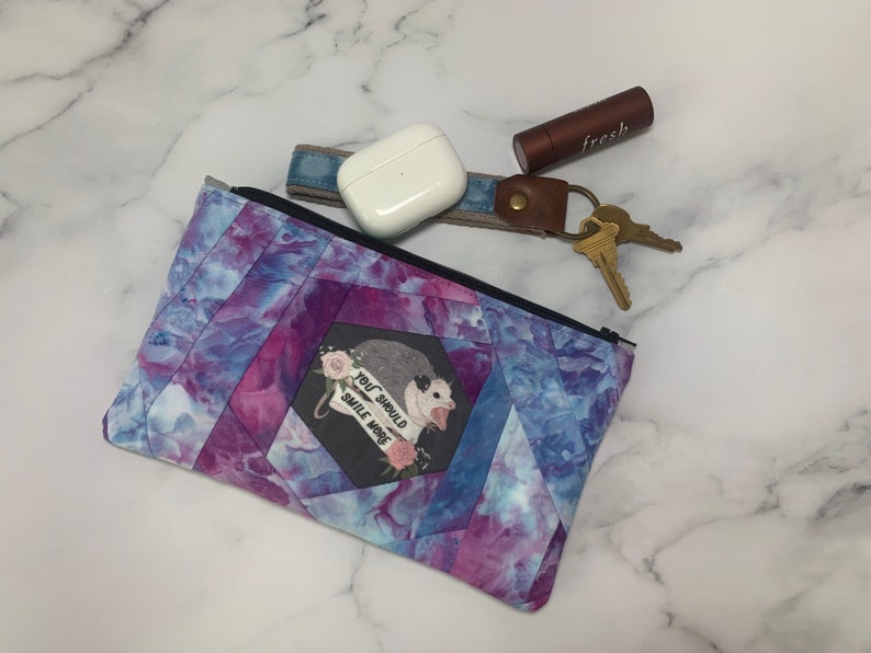 May include: A quilted fabric pouch with a black zipper, featuring a tie-dye pattern in shades of blue, purple, and pink. The pouch has an image of an opossum with the text "YOU SHOULD SMILE MORE". Beside the pouch are keys, a lipstick, and a pair of white wireless earbuds.