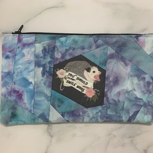 May include: A rectangular zippered pouch with a blue and purple watercolor pattern. The pouch features a cartoon opossum with the text "YOU SHOULD SMILE MORE" on a black background. The zipper is black.