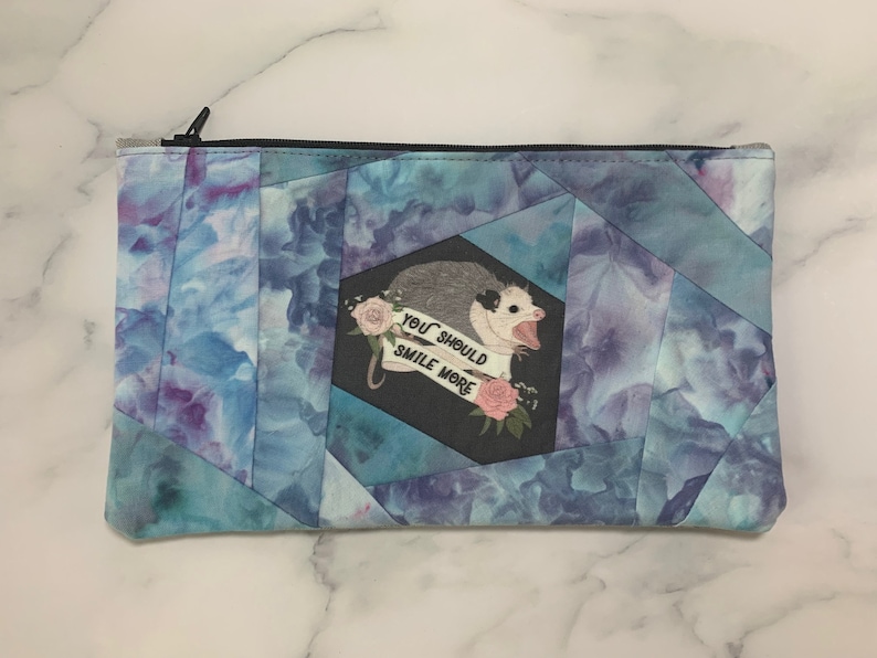May include: A rectangular zippered pouch with a blue and purple watercolor pattern. The pouch features a cartoon opossum with the text "YOU SHOULD SMILE MORE" on a black background. The zipper is black.