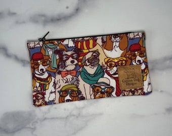 May include: A rectangular zippered pouch with a colorful pattern of dogs wearing glasses, scarves, and hats. The pouch has a black zipper and a small brown tag that reads "Curious Dog Designs."