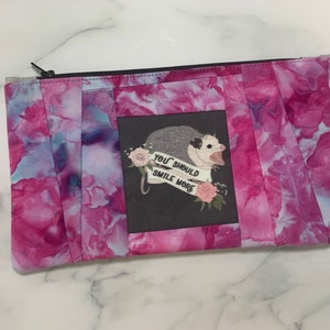 May include: A rectangular zippered pouch with a vibrant pink and blue watercolor pattern. The center features a gray square with an opossum illustration and the text "YOU SHOULD SMILE MORE". The pouch has a black zipper.