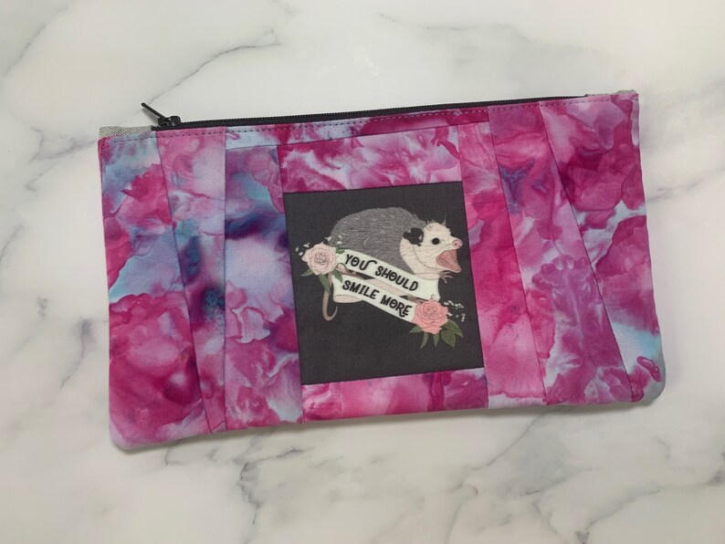 May include: A rectangular zippered pouch with a vibrant pink and blue watercolor pattern. The center features a gray square with an opossum illustration and the text "YOU SHOULD SMILE MORE". The pouch has a black zipper.