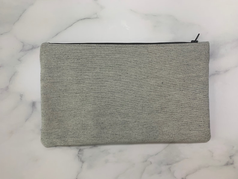 May include: A rectangular, light gray zippered pouch made of a textured fabric. The zipper is black, and the pouch is closed. The pouch is a neutral color and could be used for various purposes.