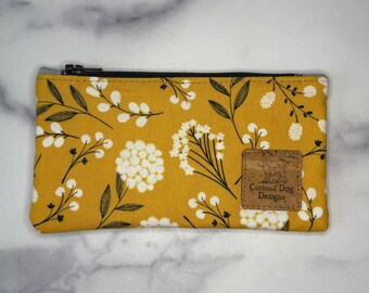 May include: A rectangular, zippered pouch with a mustard yellow floral pattern. The design features white flowers and black leaves. A brown leather tag with the text "Curious Dog Designs" is sewn on the front.