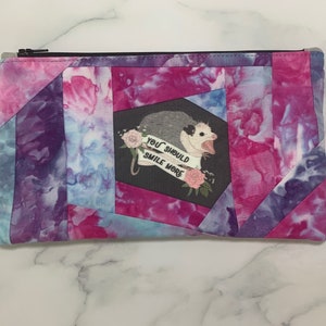 May include: A rectangular zippered pouch with a tie-dye pattern in shades of pink, purple, and blue. The pouch features a graphic of an opossum with the text "YOU SHOULD SMILE MORE". The design is set against a black background.