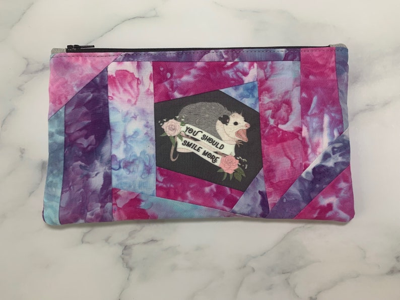 May include: A rectangular zippered pouch with a tie-dye pattern in shades of pink, purple, and blue. The pouch features a graphic of an opossum with the text "YOU SHOULD SMILE MORE". The design is set against a black background.