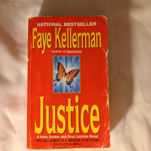 May include: A paperback book titled "Justice" by Faye Kellerman. The cover is predominantly red with the title in large yellow letters. A butterfly graphic is centered on the cover. The book is a "National Bestseller."