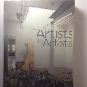 May include: A book cover with the title "Artists to Artists" in a white room with a wooden sculpture and paint supplies on a table.