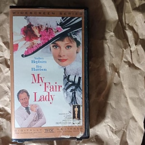 May include: A VHS tape of the movie "My Fair Lady" featuring Audrey Hepburn and Rex Harrison. The cover art includes a portrait of Hepburn in a hat, with the film's title in red and the text "Widescreen Series". The tape is in a black case.