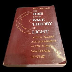 May include: A hardcover book titled "The Rise of the Wave Theory of Light" by Jed Z. Buchwald. The cover is a deep red with gold lettering and a stylized design. The book's subtitle is "Optical Theory and Experiment in the Early Nineteenth Century."