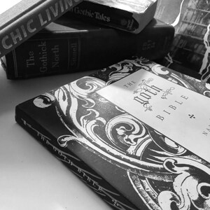 May include: A stack of vintage books in black and white. The top book is titled "The Goth Bible" with ornate white designs. Other titles include "Chic Living" and "The Gothick North". The books are arranged on a light surface.