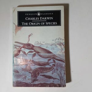 May include: A vintage copy of "The Origin of Species" by Charles Darwin, published by Penguin Classics. The book cover features an illustration of prehistoric marine reptiles in shades of brown, blue, and white. The title and author's name are in black text.