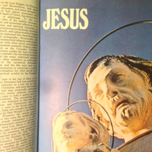 May include: A close-up of a sculpture of Jesus with a halo, against a blue sky background. The sculpture is made of a light-colored stone and shows Jesus with a beard and long hair. The word "JESUS" is written in large, yellow letters at the top of the image.