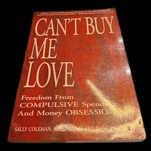 May include: A hardcover book titled "CAN'T BUY ME LOVE" with the subtitle "Freedom From Compulsive Spending And Money Obsession." The cover is red with white lettering. The author's name is visible at the bottom.