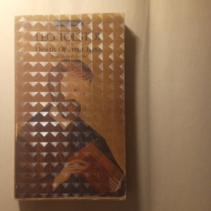 May include: A vintage paperback book cover featuring a portrait of Leo Tolstoy, the author of "The Death of Ivan Ilyich and Other Stories". The cover is a yellowed brown color with a silver geometric pattern.