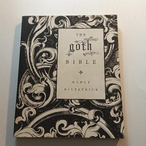 May include: A white ceramic mug with a black handle and the text "But First, Coffee" in a bold, black font. The mug is set against a blurred background, suggesting a cozy setting.