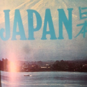May include: A vintage travel poster with a blue sky and a mountain range in the background. The word "JAPAN" is written in large, blue letters across the top of the poster. The word "日本" is written in smaller, black letters to the right of the word "JAPAN".