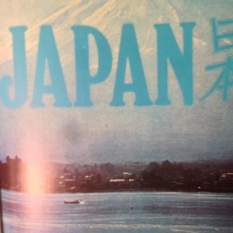 May include: A vintage travel poster with a blue sky and a mountain range in the background. The word "JAPAN" is written in large, blue letters across the top of the poster. The word "日本" is written in smaller, black letters to the right of the word "JAPAN".