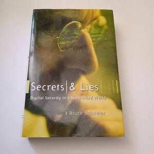 May include: A book titled "Secrets & Lies: Digital Security in a Networked World" by Bruce Schneier. The cover features a close-up of a person's face with glasses, partially obscured by a yellow and green background.