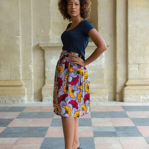 May include: A woman with curly brown hair wearing a dark blue short-sleeved shirt and a colorful floral skirt. She is standing on a black and white checkered floor.