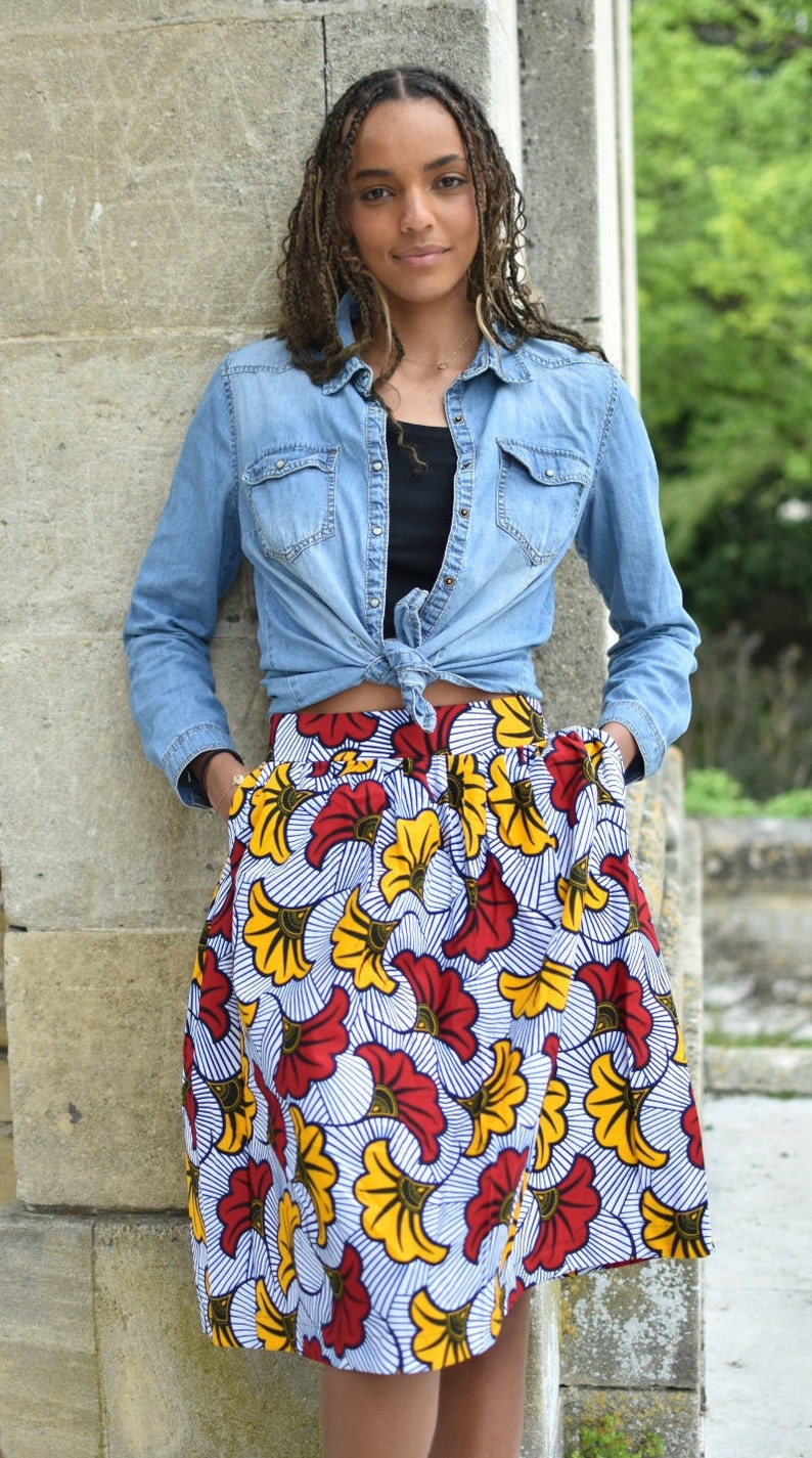 May include: A woman wearing a blue denim shirt tied at the waist over a black top and a colorful floral skirt with red, yellow, and white flowers.