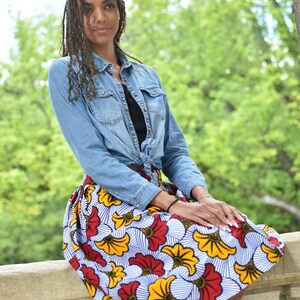 May include: A woman wearing a denim shirt tied at the waist over a black top and a colorful skirt with a floral pattern. The skirt has red, yellow, and white flowers on a blue and white striped background.