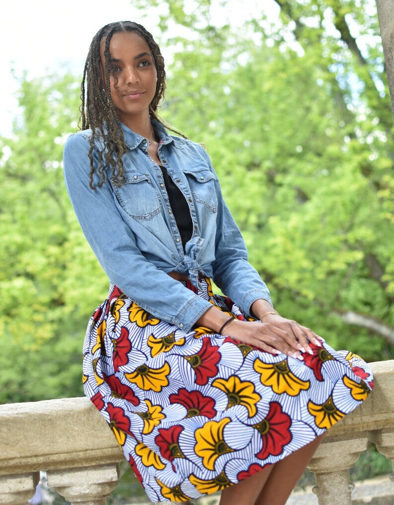 May include: A woman wearing a denim shirt tied at the waist over a black top and a colorful skirt with a floral pattern. The skirt has red, yellow, and white flowers on a blue and white striped background.