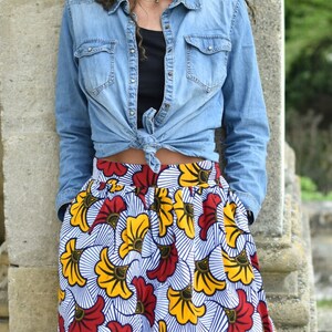 May include: A woman wearing a blue denim shirt tied at the waist over a black tank top and a colorful skirt with a white background and red, yellow, and blue floral print.