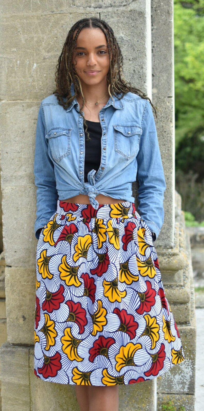 May include: A woman wearing a blue denim shirt tied at the waist over a black tank top and a colorful skirt with a white background and red, yellow, and blue floral print.
